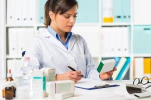Female pharmacist sat at desk writing notes with medicine boxes in background.