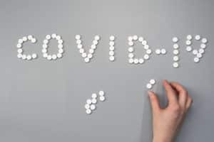 The words Covid-19 spelled out with pills, a hand in the foreground placing some pills.