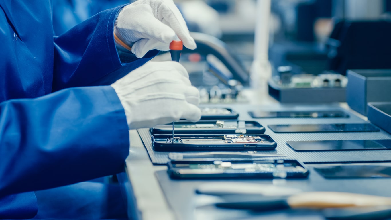 Close Up of Electronics Factory Worker in Blue Work Coat and Protective Glasses Assembling Smartphones with Screwdriver. High Tech Factory Facility with more Employees in the Background.
